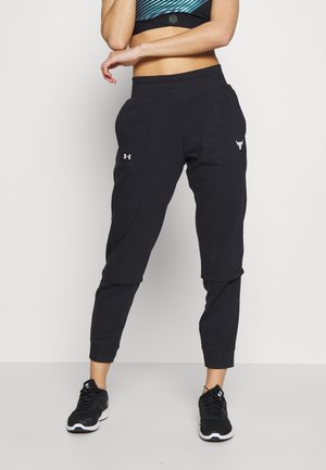 Kobieta ubrana w czarne, warstwowe spodnie dresowe Under Armour i czarne buty sportowe, stojąca na jednolitym białym tle.