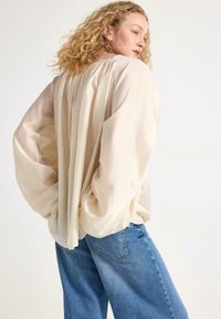 Ljus beige, flärdig blus med veckad rygg och voluminösa ärmar, tillsammans med högmidjade blå denimjeans. Slät textur, avslappnad passform.