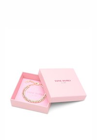 Guldtonet armbånd med glitrende perler, præsenteret i en lyserød boks med blødt indre. Boksen har teksten "TONE DAMLI by A&C" på toppen.