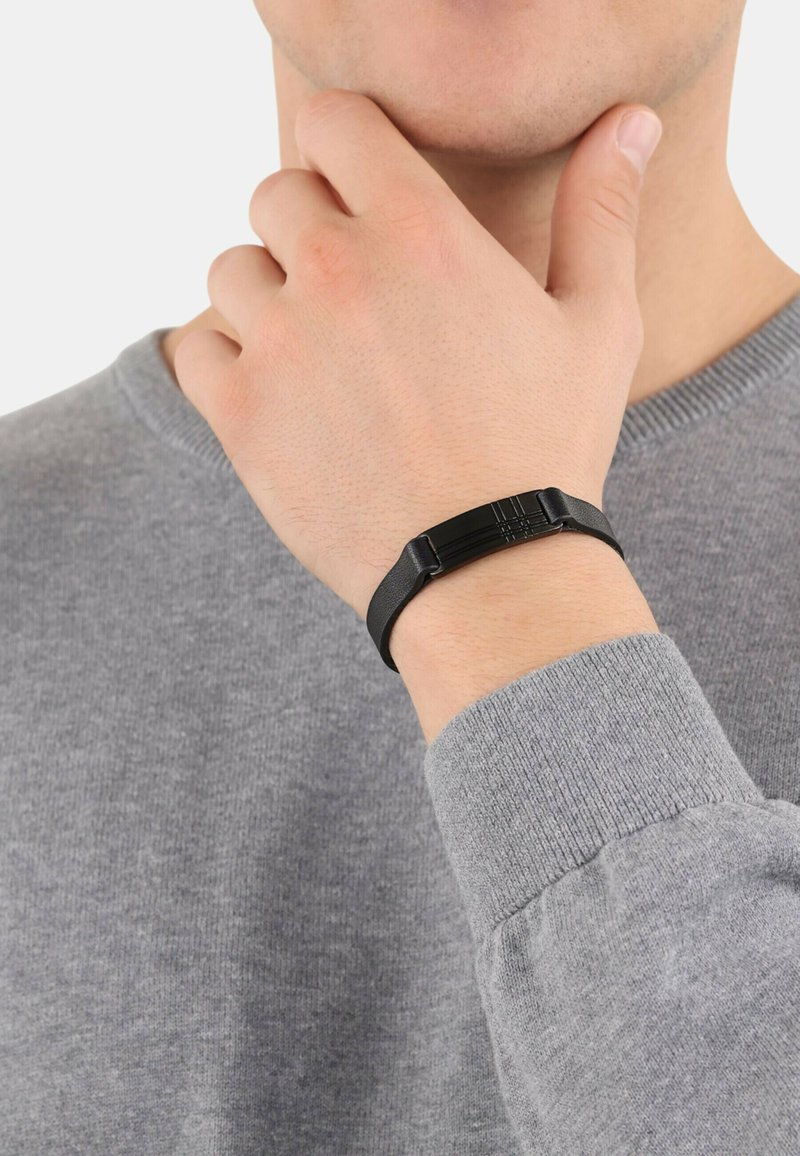 Pulsera de goma negra con un cierre angular texturizado, llevada en una muñeca de piel clara, sobre un fondo de suéter gris.