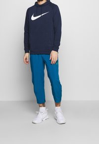 Uomo che indossa un felpa Nike blu navy, pantaloni turchesi, sneakers bianche e calzini bianchi, in piedi su uno sfondo grigio chiaro uniforme.