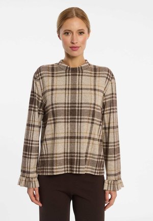 Vrouw die een beige-bruine geruite top met lange mouwen en gerimpelde manchetten draagt en donkerbruine broek, staand tegen een witte achtergrond.