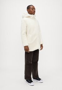 Veste à capuche blanche et fluffy avec une fermeture éclair intégrale et des poches latérales, associée à un pantalon en velours côtelé marron foncé et des baskets noires.