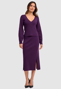Diep paarse V-hals, lange mouwen, geribbelde gebreide top en bijpassende knie-lange rok met zijssplit, gecombineerd met zwarte hoge hakken.