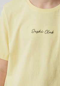 Geel katoenen t-shirt met korte mouwen en een ronde hals, met "Sushi Club" in zwarte cursieve letters geborduurd op de borst.