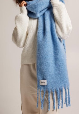 Personne portant un pull blanc côtelé et un pantalon beige, tenant une grande écharpe bleue claire et duveteuse avec des extrémités frangées et une étiquette de tissu visible.