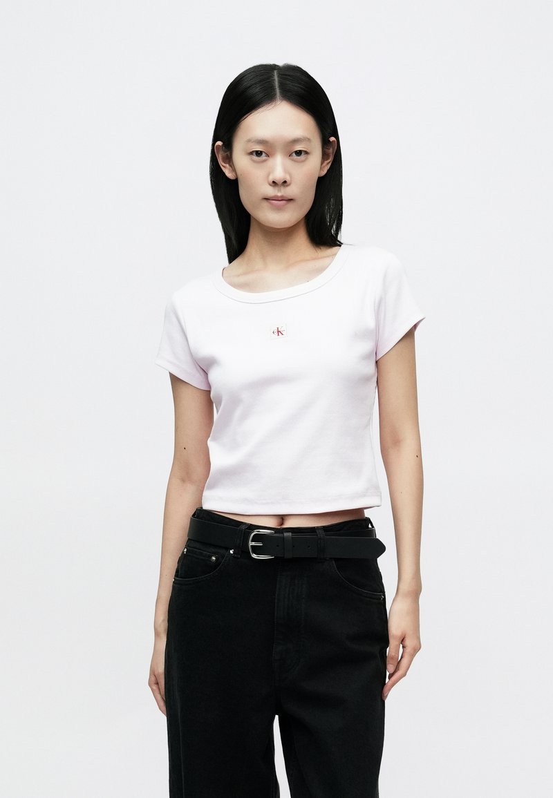 Light pink cropped t-shirt with short sleeves, ribbed texture, and a small white logo on the chest, paired with high-waisted black trousers and a belt.