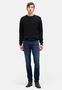 Czarny sweter z dzianiny z okrągłym dekoltem, zestawiony z ciemnoniebieskimi dżinsami i czarnymi botkami, stojąca na jednolitym białym tle.