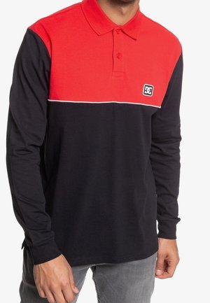 Mężczyzna noszący polo z długim rękawem, z czerwoną górną częścią, czarną dolną i małym białym logo na piersi.