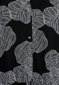 Schwarze Bluse mit großen weißen blattartigen Mustern, aus strukturiertem Stoff und mit knöpfbarem Vorderteil. Die Knöpfe sind grau und rund.