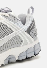 Chaussure de sport grise avec une tige en mesh et synthétique, des accents texturés, des bandes blanches et une semelle rembourrée avec des pods segmentés.