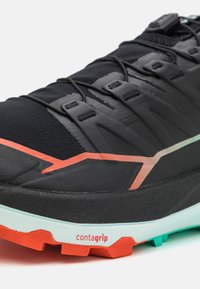 Zapatilla deportiva negra con material texturizado, presenta un acento rojo y blanco, una parte superior transpirable y una suela multicolor marcada "contagrip".