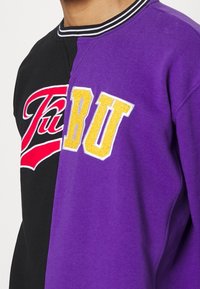 Czarna i fioletowa bluza z rozdzielonym wzorem, haftowanymi logo w kolorach czerwonym i żółtym. Posiada prążkowany kołnierz oraz miękką, polarową fakturę.