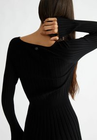 Pull en tricot côtelé noir avec des manches longues, un décolleté bardot et un accent à l'épaule mettant en avant un petit logo métallique.