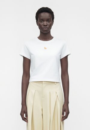 Ženska stoji obrnjena proti spredaj, nosi bel kratki top z majhnim oranžnim lisjim logotipom in bež visokim pasom, gubane hlače.