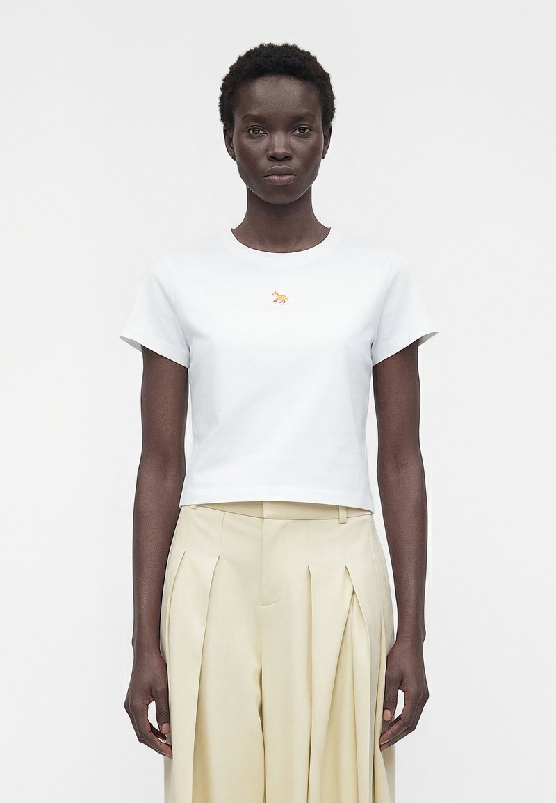 Γυναίκα που στέκεται μπροστά φοράει ένα κοντομάνικο λευκό crop top με μικρό πορτοκαλί λογότυπο αλεπούς και μπεζ υφασμάτινη φούστα με πιέτες και ψηλή μέση.