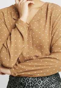 Blusa marrone a pois con scollo a V, maniche lunghe e chiusura a bottone ai polsini, abbinata a una gonna verde a fantasia.