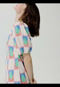 Žena sa smeđom kosom do ramena, odjevena u haljinu kratkih rukava s višebojnim kariranim i zelenim apstraktnim uzorcima, okrenuta u stranu.