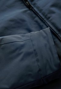 Material de culoare bleumarin, cu o textură netedă, dotat cu închidere cu fermoar și detalii la mansete. Materialul pare ușor și rezistent la apă.