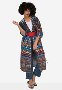 Monivärinen kimono, jossa on sahalaitakuvioita ja kukkakuoseja, pitkät hihat, avonainen etuosa, kiinnitetty punaisella vyöllä, joka on puettu valkoisen paidan ja farkkujen päälle.