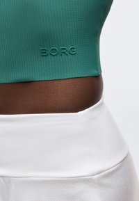 Κοντινό πλάνο της μέσης ενός ατόμου με έναν τιρκουάζ ριμπ πλεκτό κοντό τοπ με την επιγραφή «BORG» και λευκά ψηλόμεσα κάτω ρούχα.