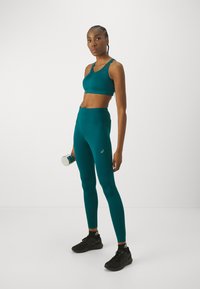 Mėlynos sportinės liemenėlės ir aukšto juosmens leggingai iš elastingos medžiagos. Modelis laiko sidabrinį hantelį, dėvėdamas juodus sportinius batelius.