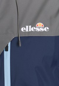 Gros plan d'une veste grise et marine avec une fermeture éclair bleu clair et un logo ellesse blanc avec un demi-cercle orange au-dessus.