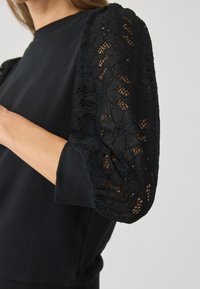 Sudadera negra con mangas de encaje abullonadas. Cuerpo de tejido suave, con intrincados detalles de encaje en las mangas, que presentan un patrón floral y un acabado texturizado.