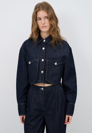 Chaqueta de mezclilla recortada en azul oscuro con botones plateados, dos bolsillos en el pecho y costuras en contraste. Combinada con pantalones de tiro alto a juego.