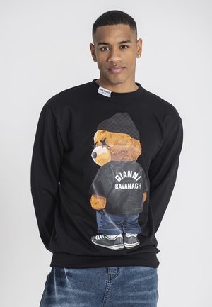 Sudadera negra con un gráfico de un oso de dibujos animados en la parte posterior, que lleva puesta una chaqueta oscura con el texto "GIANNI KAVANAGH" y unos jeans de color azul claro.