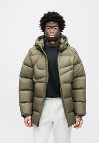 Oljčno zelena puhasta jakna s kapuco, prešita zasnova, z zadrgo spredaj in nastavljivimi zaponkami na manšetah; oblečena preko črne majice in svetlo sivih hlač.