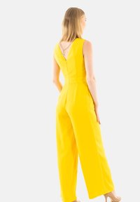 Tuta gialla con gambale largo, scollatura a V profonda sul retro e finitura in tessuto liscia. Presenta un accento a catena sulla scollatura posteriore.