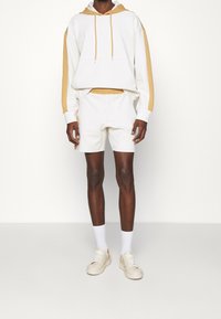 Vit huvtröja med beige sidostråkar, kombinerad med vita shorts. Bärs med vita strumpor och beige sneakers.