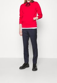 Czerwona bluza z kapturem i czarnymi sznurkami, noszona na białej koszulce; w zestawieniu z granatowymi spodniami slim-fit i czarnymi sznurowanymi butami.