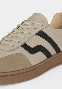 Basket beige et sable avec bandes latérales noires, lacets beiges, semelle en gomme et marquage doré "GANT" sur le côté et la languette.