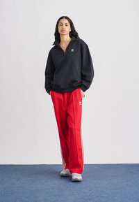 Svart tröja med dragkedja och vitt logo, kombinerad med röda träningsbyxor med vita ränder. Grå sneakers fulländar outfiten.