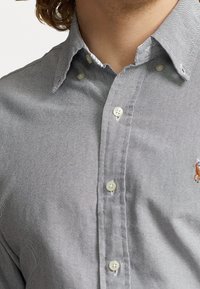 Camicia grigia con bottoni, realizzata in tessuto texturizzato. Presenta un colletto a punta, piccoli bottoni e una patch con logo sul petto.
