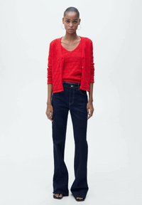 Rode gebreide cardigan en bijpassend topje, gecombineerd met donkerblauwe uitlopende jeans. Het ensemble heeft een gestructureerd patroon en een getailleerd ontwerp.