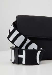 Borsa a tracolla nera con una finitura liscia e opaca. Presenta una robusta tracolla in tessuto a strisce bianche e nere con una fibbia metallica.
