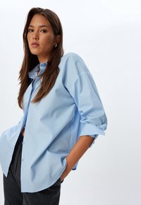 Camicia oversize azzurra realizzata in tessuto liscio, con abbottonatura frontale, maniche arrotolate e orlo curvo, abbinata a pantaloni a righe.