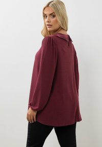 Kvinna med långt blont hår som bär en ribbad burgundyröd långärmad tröja med en rutig krage, som ser över axeln mot en enfärgad bakgrund.