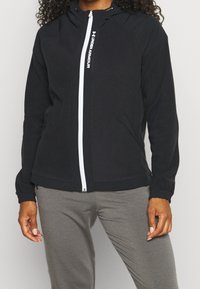Giacca nera con zip realizzata in tessuto morbido, dotata di cappuccio, zip bianca e dettaglio con logo. Presenta polsini elasticizzati e tasche laterali.
