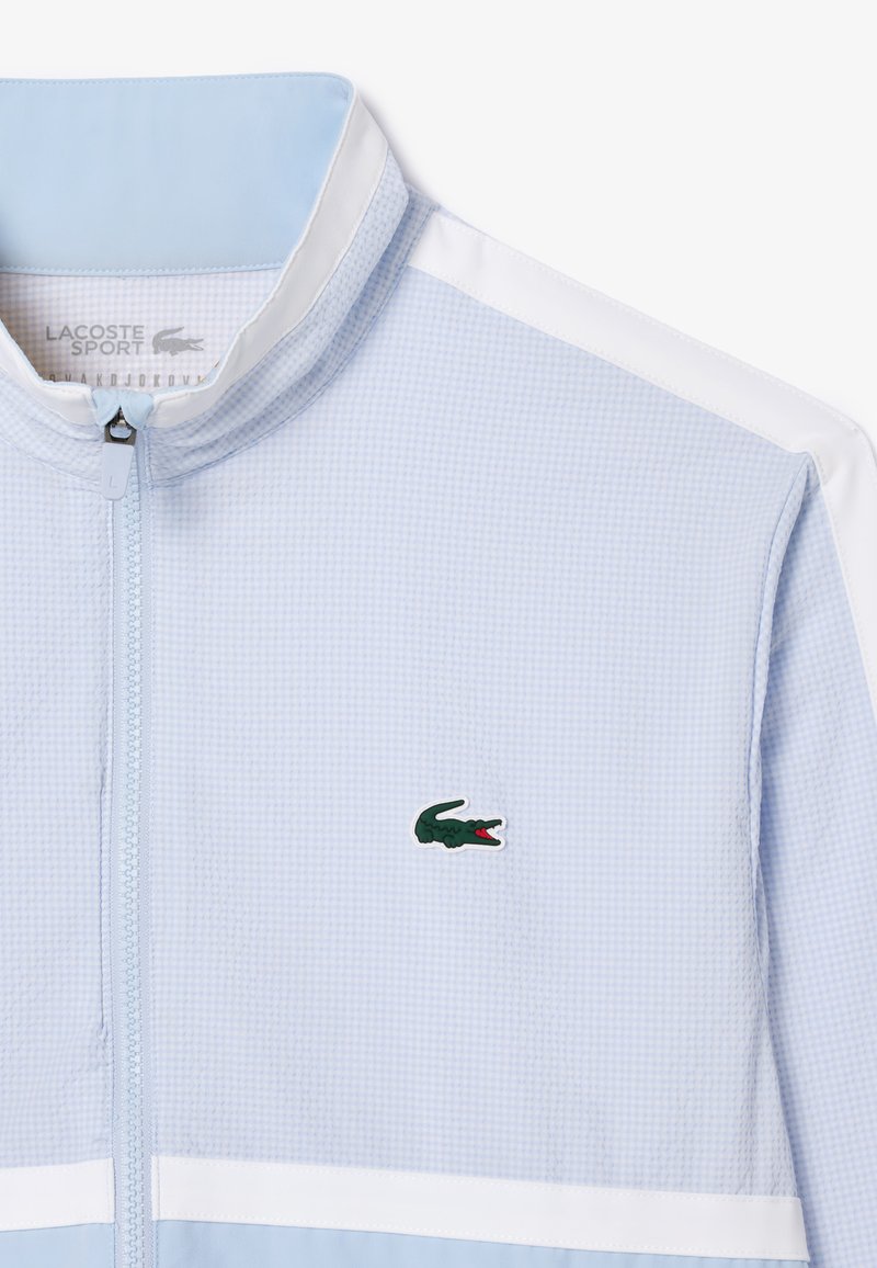 Veste zippée bleu clair avec un motif texturé, des rayures blanches et un logo de crocodile vert sur la poitrine.