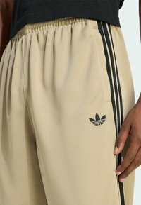 Béžové tepláky Adidas s čiernymi pruhmi a logom, nosené osobou s tmavou pokožkou, ruka spočíva pri bočnom šve.