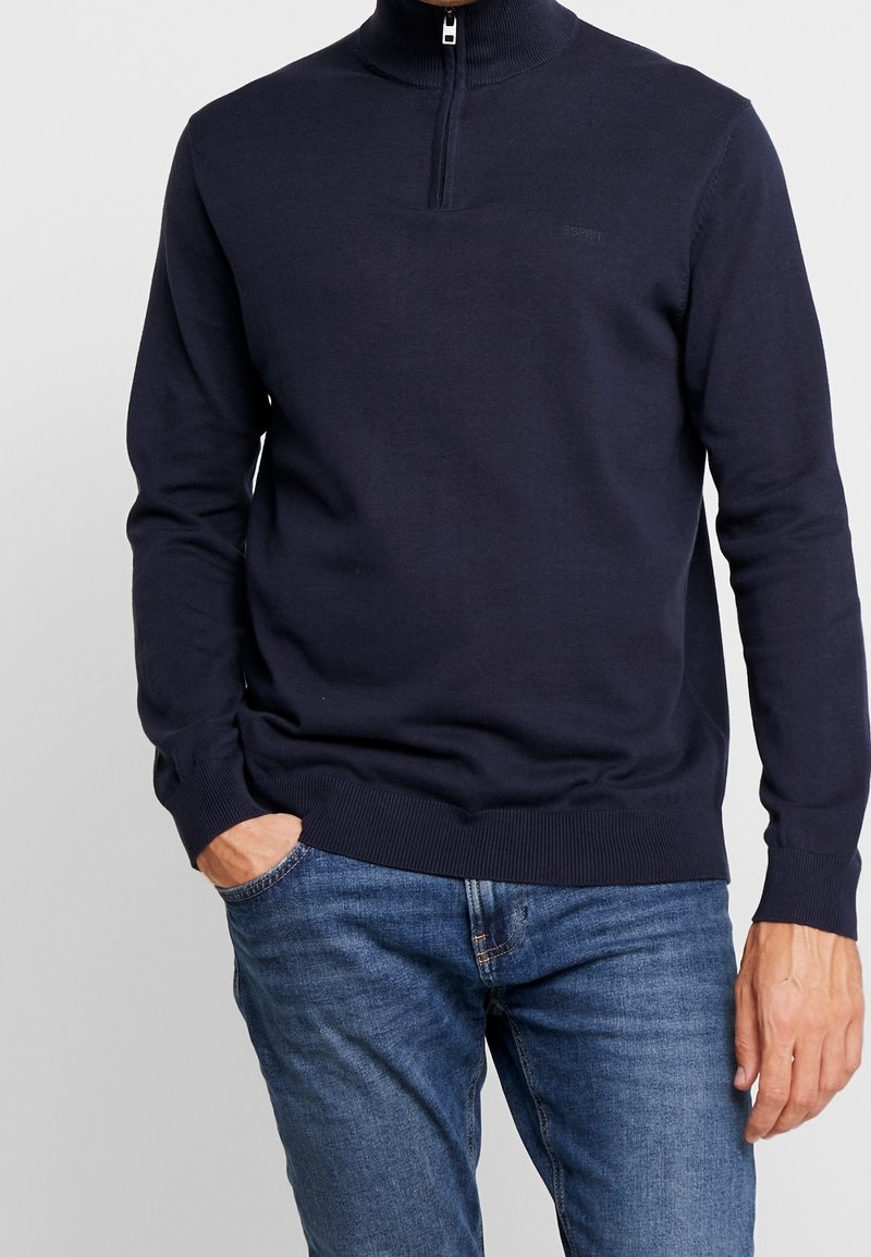 Homme portant un pull bleu marine à fermeture éclair quart et un jean bleu avec la main gauche dans la poche, sur fond uni.
