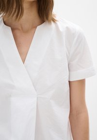 Blusa bianca a maniche corte con scollo a V e tessuto strutturato. Presenta un design semplice e sovrapposto con un accento di tasca laterale.