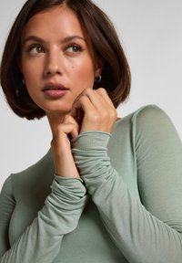 Top verde chiaro a maniche lunghe con dettaglio arricciato ai polsini. Tessuto morbido e sottile con leggero lucido. Le mani della modella sono incrociate vicino al mento.