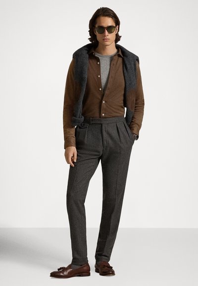 Vyras, dėvintis akinius nuo saulės, rudos spalvos marškinį ant pilko megztinio, tamsiai pilkos kelnes, rudus odinius laisvalaikio batelius ir tamsiai pilką megztinį per petį.