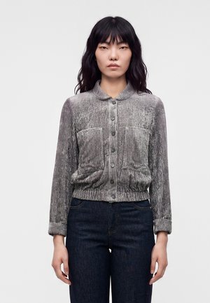 Kvinde med langt sort hår iført en grå tekstureret jakke med knapper og mørkeblå jeans, stående mod en ensfarvet baggrund.