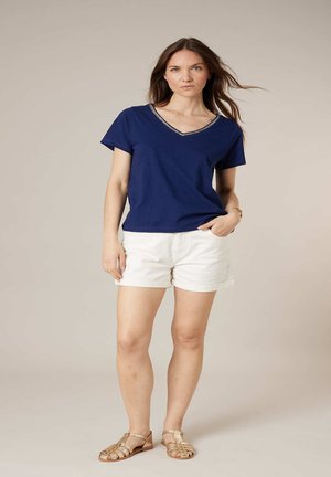 Femme debout, main dans la poche, portant un haut bleu marine à manches courtes, un short blanc et des sandales beige, sur un fond uni.